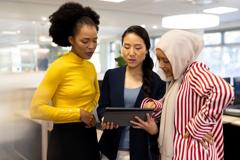 Três mulheres conversam enquanto analisam algo em um tablet, demonstrando a importância do networking.