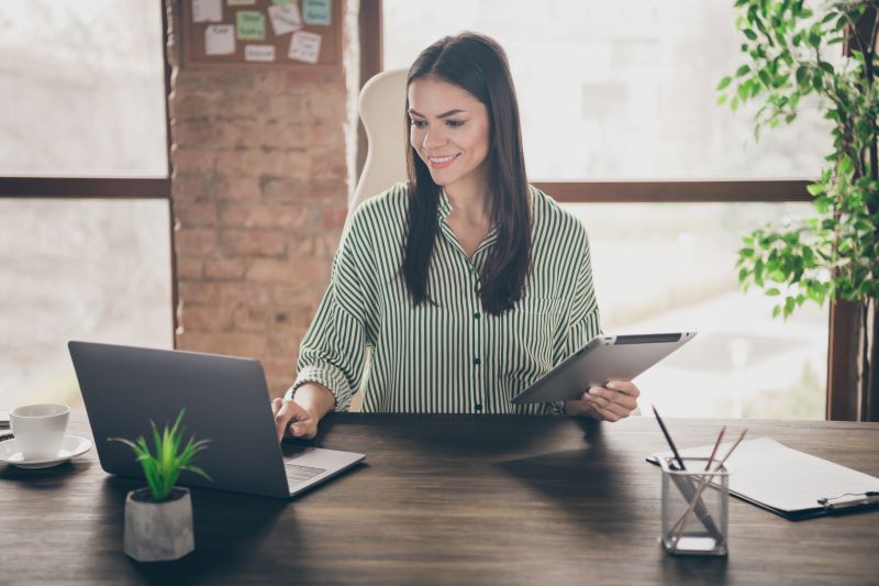 Uma mulher trabalha em seu escritório utilizando um notebook e um tablet. 