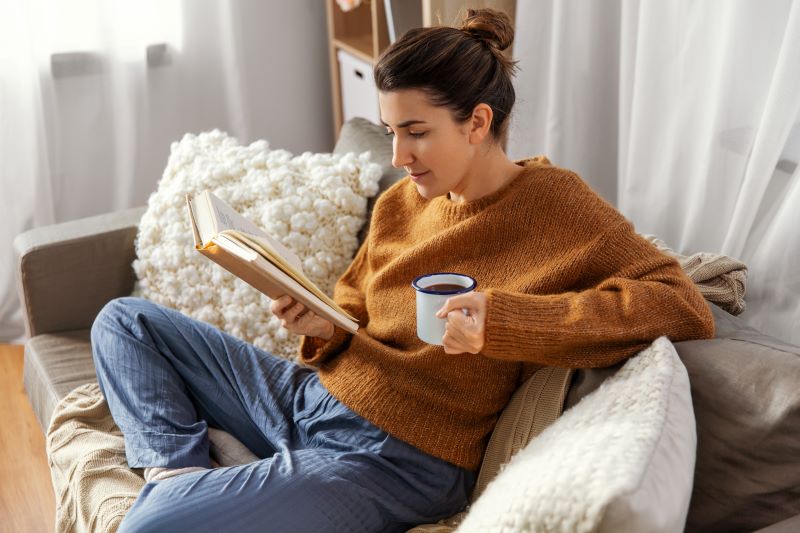 Mulher lendo um dos livros para desenvolvimento pessoal que possui, no sofá de casa e com uma caneca de café.