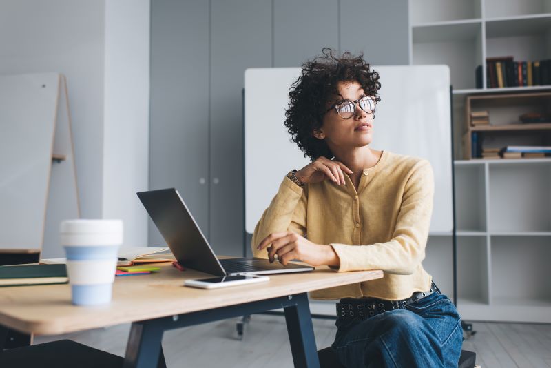 Quero empreender mas não tenho ideias: mulher pensativa em sala de reunião, sozinha, olhando para a janela enquanto usa notebook.