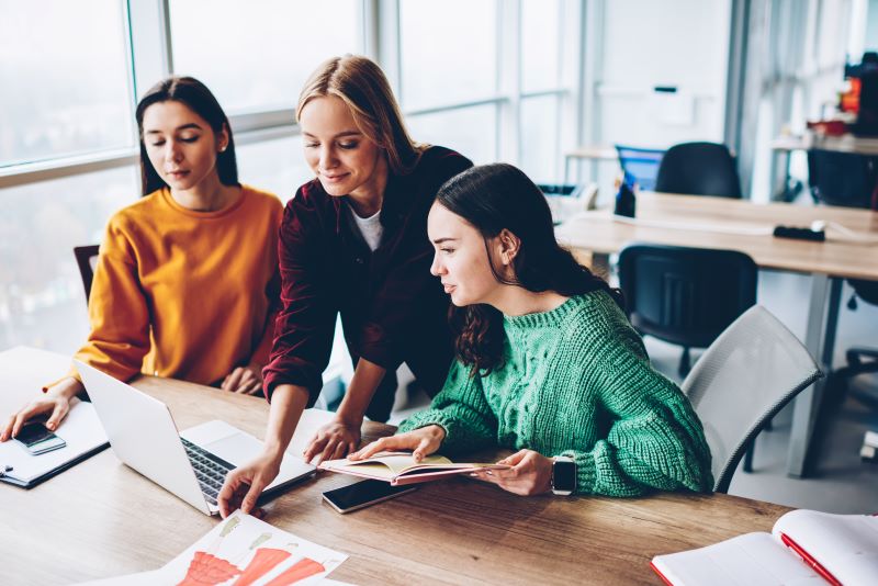 Três mulheres engajadas em trabalho colaborativo em um coworking, analisando croquis de moda com ajuda de notebook.