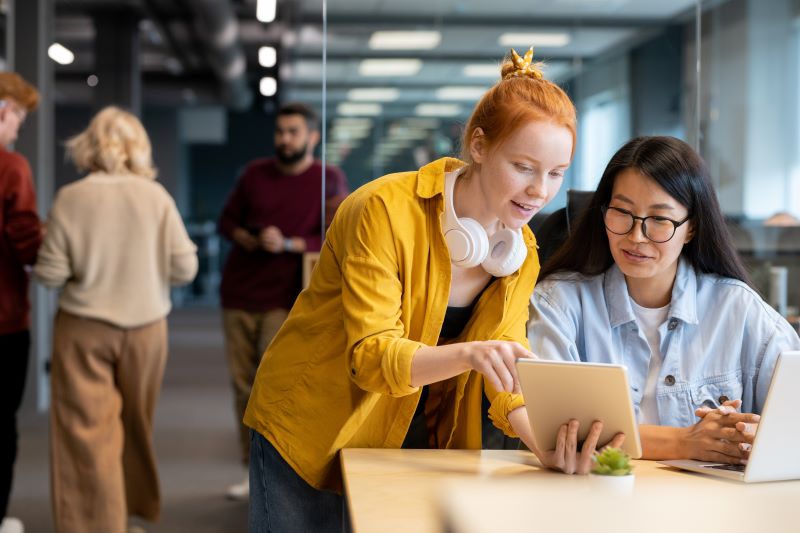 Mulheres em coworking falando sobre negócios que faltam no Brasil, pesquisando em tablet.