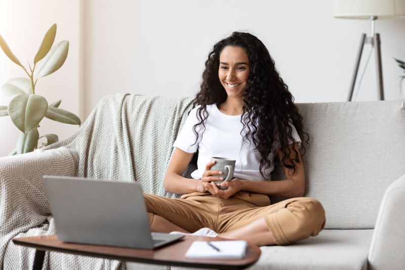 Filmes para empreendedores​: mulher assistindo filme no notebook sentada no sofá de casa com caneca na mão